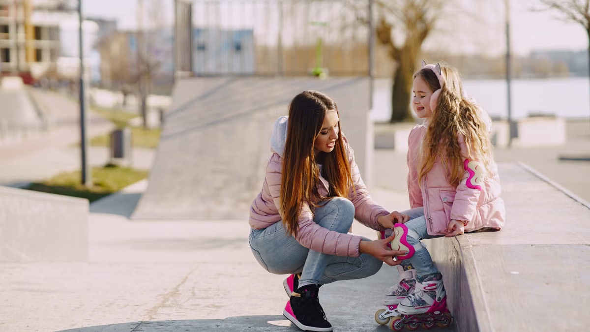 Cheerful young woman adjusting knee pads for little girl during active sport and game outside in city while spending time together