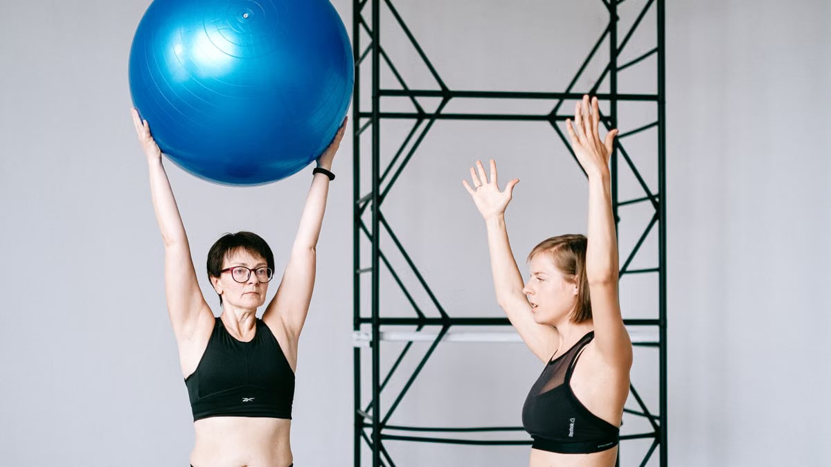 Two women exercising in a gym with a blue balance ball, promoting fitness and health.